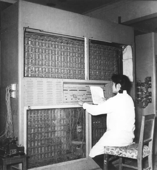 操作人员手持记录本、指向控制开关；完整竖构图保留人物与身后机柜的关系。